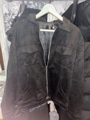 Svart mockajacka med dragkedja - Snygg svart mockajacka med klassisk krage och silverfärgad dragkedja framtill. Jackan har två bröstfickor med lock och två stora fickor nertill. Perfekt för dig som gillar stilren och enkel design med en modern touch.