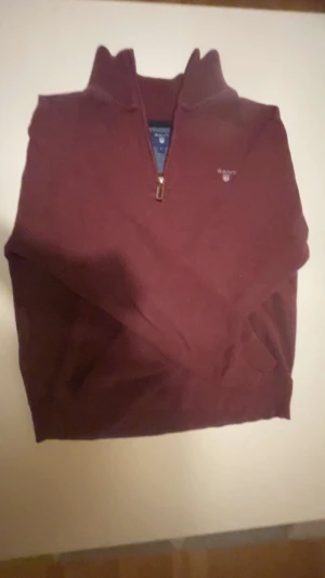 Vinröd half zip tröja från GANT - Vinröd stickad tröja från GANT med halv dragkedja och broderad logga på bröstet. Tröjan har ribbad krage och är i klassisk modell, perfekt för lager-på-lager. Materialet är mjukt och känns lyxigt mot huden.