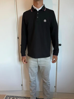 Svart långärmad Moncler skjorta - Svart långärmad skjorta från Moncler med klassisk krage och knappar framtill. Diskret Moncler-logga på bröstet och snygga rödvita detaljer på kragen och ärmsluten. Skjortan är i mjuk bomull och har en normal passform som sitter skönt.