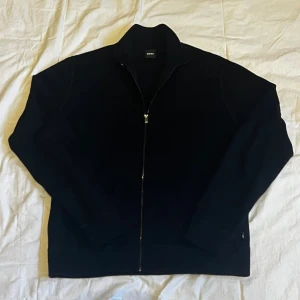 Svart kofta med dragkedja från BOSS - Svart kofta från BOSS med hel dragkedja framtill och hög krage. Tröjan är långärmad och har en stilren, klassisk look med ribbade detaljer vid kragen och muddar. Perfekt för dig som gillar enkel och snygg design.