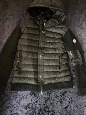Svart Moncler pufferjacka med huva - Svart pufferjacka från Moncler med huva, ribbstickade ärmar och nederkant. Jackan har quiltad kropp i syntetmaterial, dragkedja framtill och fickor med dragkedja. Moncler-logga på ärmen och extra ficka med dragkedja på vänster arm. Perfekt för kalla dagar.