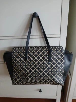 By Malene Birger  - Stilren axelväska med svart och beige geometriskt mönster. Väskan har långa svarta handtag, sidor i svart skinn och en stor dragkedja upptill. Insidan är rymlig med en innerficka. Perfekt för dig som vill ha plats för allt och samtidigt se snygg ut.