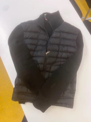 Svart moncler cardigan  - Svart pufferjacka från Moncler med quiltad framsida och släta ärmar. Jackan har två dragkedjeförsedda fickor, hög krage och Moncler-logga på ena ärmen. Materialet är en mix av polyester och stickad textil, perfekt för kyliga dagar.