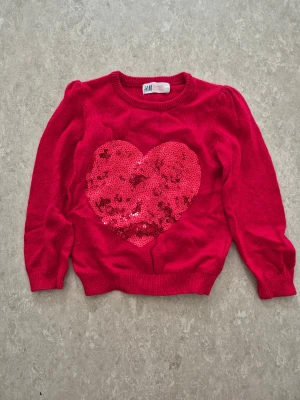 Stickad tröja - Snygg röd stickad tröja från H&M med ett stort hjärta i röda paljetter på bröstet. Tröjan har rund hals, puffade axlar och långa ärmar. Perfekt för dig som gillar färg och glitter! Materialet är mjukt och bekvämt.