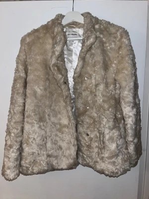 Beige pälsjacka från KappAhl - Säljer en fluffig och mjuk beige pälsjacka från KappAhl i storlek 170. Jackan har silkeslen insida i ljusgrått och stängs med diskreta knappar framtill. Perfekt för dig som vill ha en cozy och stylish look under kyliga dagar.