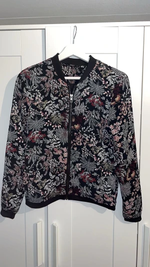 Blommig bomberjacka från ONLY - Säljer en svart bomberjacka från ONLY med blommigt mönster i vitt, rosa, rött och beige. Jackan har dragkedja framtill, ribbade muddar och lång ärm. Perfekt för dig som vill ha en cool och unik look med mycket detaljer.