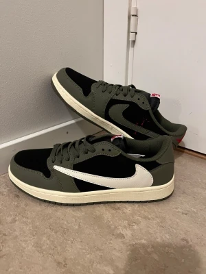 Nike Air Jordan 1 Low Travis Scott - Säljer ett par Nike Air Jordan 1 Low Travis Scott med olivgröna och svarta paneler, vit omvänd swoosh och röda broderade detaljer på hälen. Skorna har platt sula, snörning och är tillverkade i skinn och syntet. Perfekt för dig som gillar streetwear och unika sneakers.