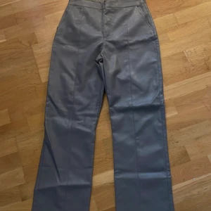 Gråa högmidjade skinnbyxor wide - Snygga gråa skinnbyxor med hög midja och raka, breda ben. Byxorna har stilrena sömmar framtill och sidofickor. Perfekta för dig som vill ha en cool och trendig look med lite edge.