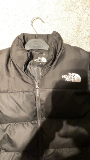 Svart dunväst från The North Face - Svart dunväst från The North Face med hög krage och två sidofickor med dragkedja. Västen har en quiltad design och logotypen i vitt på bröstet. Perfekt för lager-på-lager och chill stil under kyliga dagar.