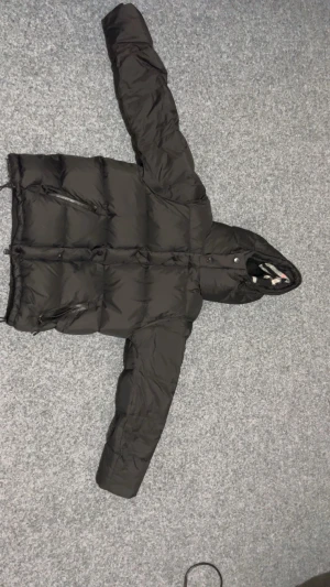 Svart pufferjacka från Burberry - Svart pufferjacka från Burberry med klassiskt rutigt foder i huvan. Jackan har dragkedja, flera fickor och justerbar nederkant med snörning. Materialet är bomull och polyester, perfekt för kalla dagar. Snygg och tidlös design med lång ärm och hög krage.