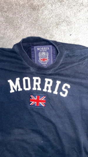 Mörkblå sweatshirt från Morris - Snygg mörkblå sweatshirt från Morris med vit broderad logga och Union Jack-flagga på bröstet. Tröjan har rund hals och långa ärmar, samt en liten patch på ärmen. Perfekt för dig som gillar klassisk brittisk stil.