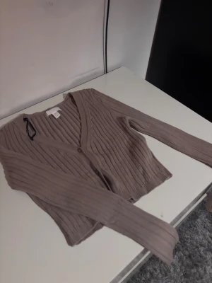 Beige ribbad croppad kofta - Säljer en beige croppad kofta från H&M med ribbad struktur och v-ringning. Koftan har långa ärmar och knäpps med två knappar framtill. Perfekt att styla med ett linne under för en chill look.