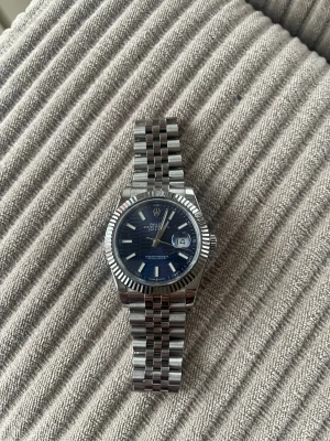 Blå  Datejust klocka i stål -  Datejust med blå urtavla och klassisk jubilee-länk i rostfritt stål. Klockan har räfflad bezel, datumfönster med cyclop-lins och tydliga indexmarkeringar. Snygg och tidlös design med silverfärgade detaljer och -logga på spännet.