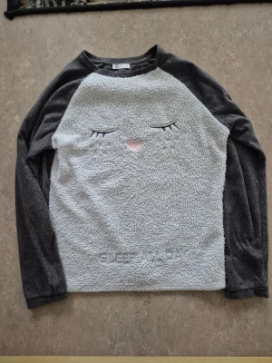 Tröja - Supermysig grå fleece-tröja från Cubus i storlek S. Framsidan är ljusgrå med broderat ansikte och texten 'SLEEP ALL DAY', medan ärmar och baksida är mörkgrå. Tillverkad i mjuk polyester, perfekt för chilliga dagar. Långärmad och loose fit.