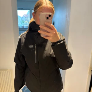 Svart skidjacka från Helly Hansen - Svart skidjacka från Helly Hansen med huva, bröstficka med dragkedja och HH-logga på både fram- och baksida. Aldrig använd. Insidan har ett mjukt foder i grått. Jackan är långärmad och har justerbara muddar med kardborre. 