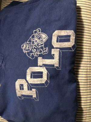 Blå hoodie från Polo Ralph Lauren - Snygg marinblå hoodie från Polo Ralph Lauren med stort tryck på bröstet och en cool maskot. Tröjan har huva, lång ärm och en klassisk känguruficka framtill. Perfekt för dig som gillar sportig och avslappnad stil.