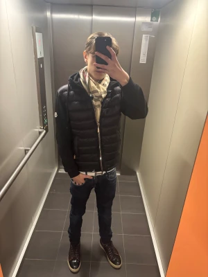 Beige rutig halsduk från Burberry - Säljer en klassisk beige halsduk från Burberry med det ikoniska rutiga mönstret i svart, vitt och rött. Halsduken har fransade kanter och broderad Burberry London-logga. Perfekt accessoar för att lyfta din outfit med lyxig känsla.