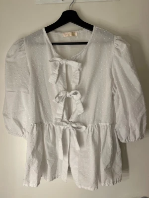 Vit blus med knyt från Chica London - Supersöt vit blus från Chica London med diskret prickigt mönster, tre knyt framtill och puffiga trekvartsärmar. Blusen har en lätt volang nedtill och är gjord i ett tunt, luftigt bomullstyg. Perfekt för en romantisk och trendig look.