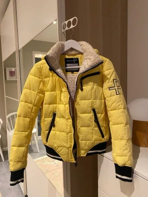Gul jacka från Pelle P - Gul jacka från Pelle P med svartvita muddar och broderad logga på ärmen och huvan. Jackan har mysigt teddyfoder, huva med snörning, flera dragkedjefickor och quiltad design. Perfekt för kalla dagar och har en sportig stil. Storlek M.