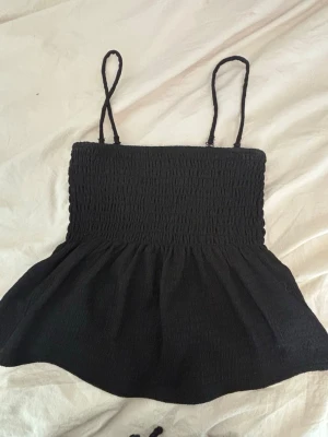 Svart smockad linnetopp med tunna band - Säljer en svart linnetopp med smockad överdel och tunna axelband. Toppen har en lätt utställd nederdel som ger en söt siluett. Perfekt för dig som gillar enkel och stilren design med en touch av volym. Passar till jeans eller kjol för en chill look. Man kan även ta av banden och få en bandeau topp.