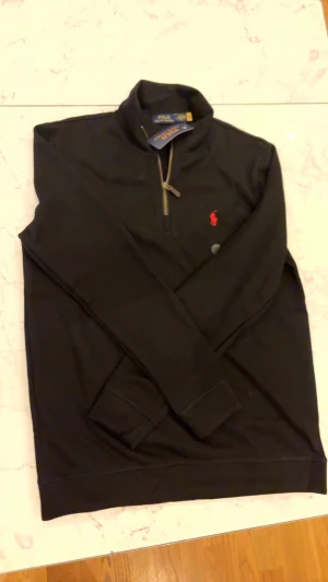 Svart half zip tröja Polo Ralph Lauren - Svart långärmad tröja från Polo Ralph Lauren med half zip och klassisk röd broderad logga på bröstet. Tröjan har ribbade muddar och är tillverkad i mjuk bomull. Perfekt för dig som gillar stilren och enkel design.