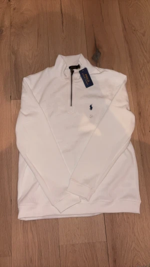Vit half zip tröja från Polo Ralph Lauren - Snygg vit långärmad tröja från Polo Ralph Lauren med half zip och klassisk blå broderad logga på bröstet. Tröjan har en hög krage och är gjord i mjuk bomull, perfekt för en clean och stilren look. Passar dig som gillar enkel design med premiumkänsla.