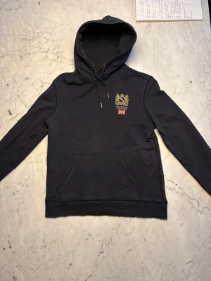 Mörkblå hoodie från Morris  - Snygg mörkblå hoodie från Morris med guldbroderat emblem och Union Jack-flagga på bröstet. Nypris ligger på cirka 1500kr, skriv om du har några frågor!