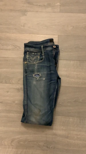Replay slim jeans med slitningar - Säljer ett par blå Replay jeans med slim passform och coola slitningar på fram- och baksidan. Jeansen har klassisk femficksdesign, låg midja och dragkedja. Materialet är jeans med snygg tvätt och detaljer som ger en avslappnad vibe. Storleken är 31/32 