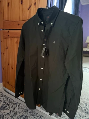 Mörkgrön skjorta från Morris XS - Stilren mörkgrön skjorta från Morris i bomull, med klassisk button down-krage och vita knappar. Broderad logga på bröstet och snygga detaljer. Perfekt för dig som gillar en clean och tidlös look. Ordinalpris 1299 kr. Skjortan är helt ny är inte använd! 