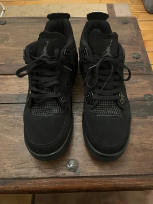 Nike Air Jordan 4 Retro Black - Säljer ett par helsvarta Nike Air Jordan 4 Retro med snygga detaljer i mesh och skinn. Klassisk siluett med snörning, Jumpman-logga på plösen och robust sula. Perfekta för dig som gillar streetwear och vill ha en ikonisk sneaker i garderoben.