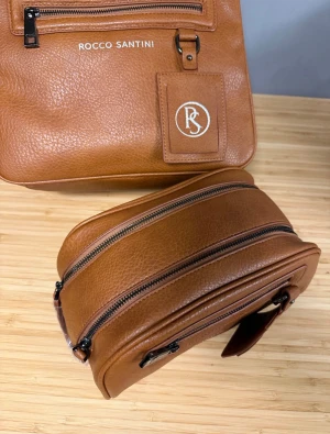 Brun ryggsäck från Rocco Santini - Snygg brun ryggsäck i skinn från Rocco Santini med stilren design och silverfärgade dragkedjor. Väskan har ett praktiskt ytterfack med dragkedja, justerbara axelremmar i brunt och svart, samt en tag med RS-logga. Perfekt för dig som vill ha en clean look.