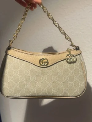 Beige axelväska  med kedja - Snygg beige axelväska med klassiskt GG-mönster och guldfärgad kedjerem. Väskan har detaljer i skinn och dragkedja upptill. Perfekt storlek för det viktigaste och ger en lyxig touch till din outfit.