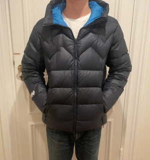 mountain works dunjacka i mörkt blå - Säljer en svart dunjacka från 8848 Altitude med blått innerfoder och huva. Jackan har quiltad design, dragkedja framtill och två sidofickor. Perfekt för kalla dagar och har en modern, sportig look.