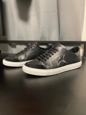 Axel ariagto skor - Snygga svarta sneakers från Axel Arigato i skinn med svarta mockadetaljer på tån och vit platt sula. På sidan finns en broderad fågel i grå och vit ton. Skorna har svarta snören och en grå detalj vid hälen. Perfekta för dig som gillar stilrena och trendiga sneakers.
