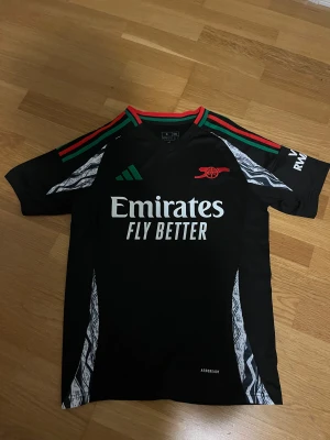 Arsenal Tröja - Svart Arsenal matchtröja från Adidas med gröna och röda detaljer på axlarna, vit logga och text på bröstet samt röd kanon-logga. Snygga mönstrade paneler i vitt och svart längs sidorna. Perfekt för dig som älskar fotboll och vill sticka ut på planen.
