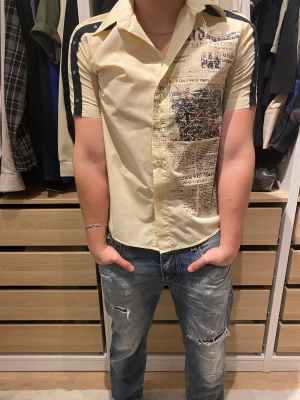 Replay skjorta med tidningsprint, L - Unik skjorta från Replay i storlek L, ljusbeige med svart tidningsmönster och orange detaljer på framsidan. Svarta ränder med nitar längs axlar och ärmar, klassisk krage och korta ärmar. Skjortan har knappar framtill och är gjord i mjukt bomullsmaterial. Defekt på en av knapparna men skjortan går fortfarande ans knäppa ordentligt.