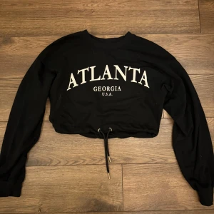 Svart croppad sweatshirt - Svart croppad sweatshirt med vit text 'ATLANTA GEORGIA U.S.A.' framtill. Tröjan har långa ärmar och snörning i nederkant för justerbar passform. Perfekt för dig som gillar streetstyle och vill ha en chill look.