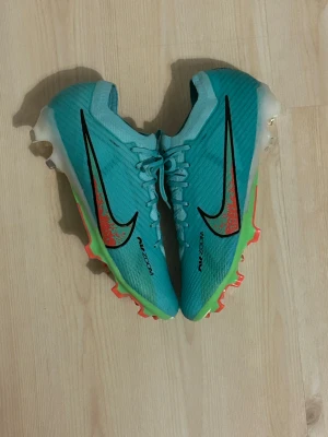 Nike Mercurial Air Zoom fotbollsskor - Nike Mercurial Air Zoom fotbollsskor i turkos/ grön med gröna och orange detaljer. Skorna har snörning, syntetmaterial och en platt sula med dobbar för gräsplan. ACC-teknologi och Vaporposite+ för extra grepp och komfort. Snygg design med mönstrade inslag och Mercurial-logga på hälen.