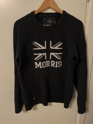 Svart stickad tröja från Morris - Marinblå stickad tröja från Morris med vit Union Jack-flagga och Morris-tryck på bröstet. Tröjan har rund halsringning, ribbade muddar och är långärmad. Perfekt för dig som gillar klassisk brittisk stil och vill ha något som sticker ut.