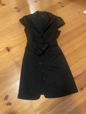 Svart kort klänning från Stockh LM - Snygg svart klänning från Stockh LM i storlek 38. Modellen är kort med knappar framtill, korta ärmar och ett bälte i midjan som ger en cool siluett. Klänningen har klassisk krage och är tillverkad i ett mjukt material som känns bekvämt mot huden.