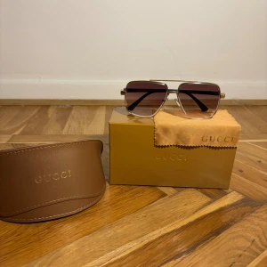 Gucci solglasögon med guldtonad ram - Snygga Gucci solglasögon med fyrkantig form och guldtonad metallram. Glasen är mörkbruna med gradient och har en cool retrovibe. Svarta skalmar och diskret Gucci-logga på linsen. Kommer med originalfodral och putsduk.