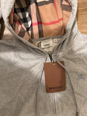 Grå hoodie från Burberry med dragkedja - Snygg grå hoodie från Burberry med klassiskt rutigt foder i huvan och broderad logga på bröstet. Tröjan har snörning i huvan, praktiska fickor och dragkedja framtill. Perfekt för dig som vill ha en stilren och lyxig look.