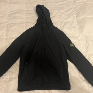 Svart vindjacka från Stone Island - Svart vindjacka från Stone Island med huva och dragkedja framtill. Jackan har två sidofickor med dragkedja och den klassiska Stone Island-loggan på vänster ärm. Materialet är syntet och passformen är normal. Perfekt för dig som gillar streetwear och clean stil.