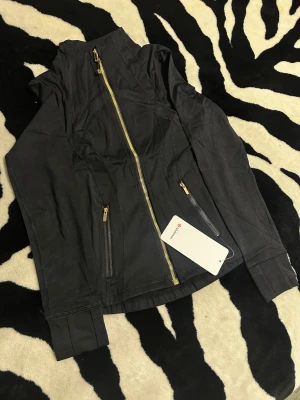 Svart Define Lululemon jacka  - Svart träningströja/jacka från Lululemon med snygga guldiga dragkedjor och detaljer. Jackan har figurnära passform, högt krage och två fickor med dragkedja framtill. Materialet är mjukt och stretchigt, tillverkat av nylon och lycra. Perfekt för träning eller chill. Detta är den korta versionen. Storlek 2