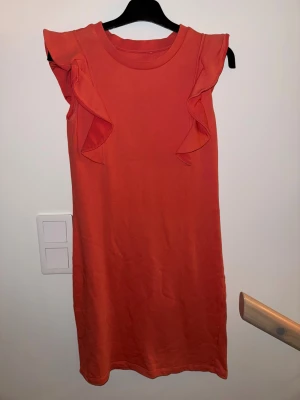 Orange ärmlös klänning med volang - Snygg orange klänning med ärmlös design och volangdetaljer vid axlarna. Enkel och rak modell som passar perfekt för varma dagar. Tillverkad i mjukt material som känns skönt mot huden.