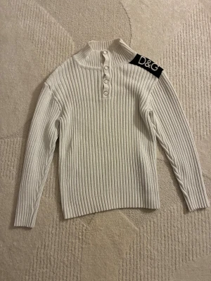 Vit ribbad polotröja från D&G - Vit ribbad polotröja från Dolce & Gabbana med silverfärgade knappar framtill och svart axelpatch med D&G-logga i pärlor. Tröjan har lång ärm och hög krage, perfekt för dig som gillar stilrena detaljer och exklusiv känsla.