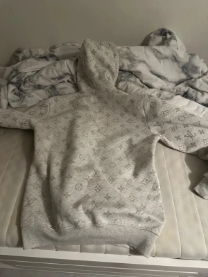 Louis Vuitton ljusgrå hoodie med monogram - Louis Vuitton hoodie i ljusgrått med ikoniskt monogrammönster över hela tröjan. Tröjan har huva med snörning, magficka och ribbade muddar. Materialet är mjukt och känns som bomull och polyester. Perfekt för dig som vill ha en clean och lyxig streetwear-look.