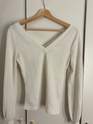 Vit ribbad blus med v-ringning - Säljer en vit blus med ribbad struktur och v-ringning både fram och bak. Toppen har långa ärmar med lätt puff och sitter snyggt figurnära. Materialet känns mjukt och stretchigt, perfekt för dig som gillar stilrena och enkla plagg med lite extra detalj.