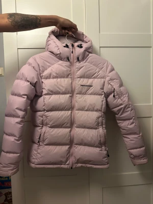 Ljusrosa Peak Performance dunjacka - Säljer en ljusrosa dunjacka från Peak Performance i storlek S. Jackan har luva, dragkedja framtill och snörning vid halsen. Den är quiltad och har praktiska fickor med dragkedja. Perfekt för kalla dagar och har en snygg, puffig look.