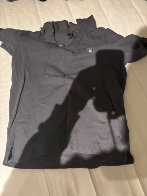 Mörkgrå pikétröja från GANT - Snygg mörkgrå pikétröja från GANT med klassisk krage och två knappar vid halsen. Diskret GANT-logga med sköld på bröstet. Tröjan är kortärmad och tillverkad i mjuk bomull som känns skön mot huden. Perfekt för en chill och stilren look.